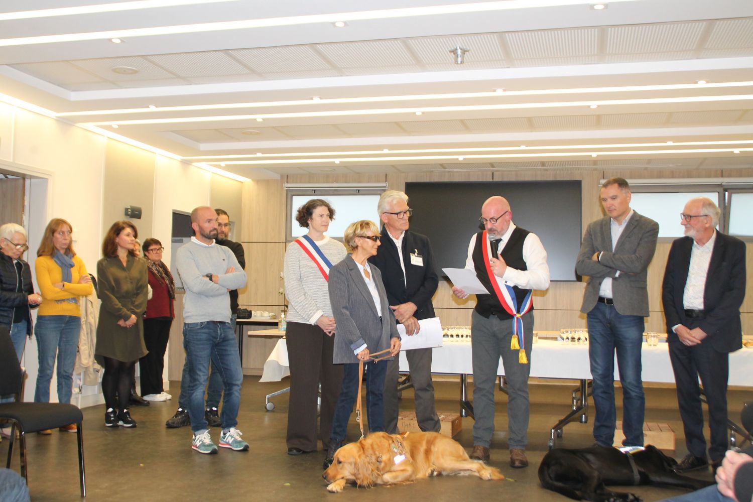 Remise officielle du chien guide Totem à sa bénéficiaire, en présence des élus et des représentants de l’association. Plusieurs personnes sont réunies autour du chien, couché au premier plan, lors de cette cérémonie organisée à Guidel.